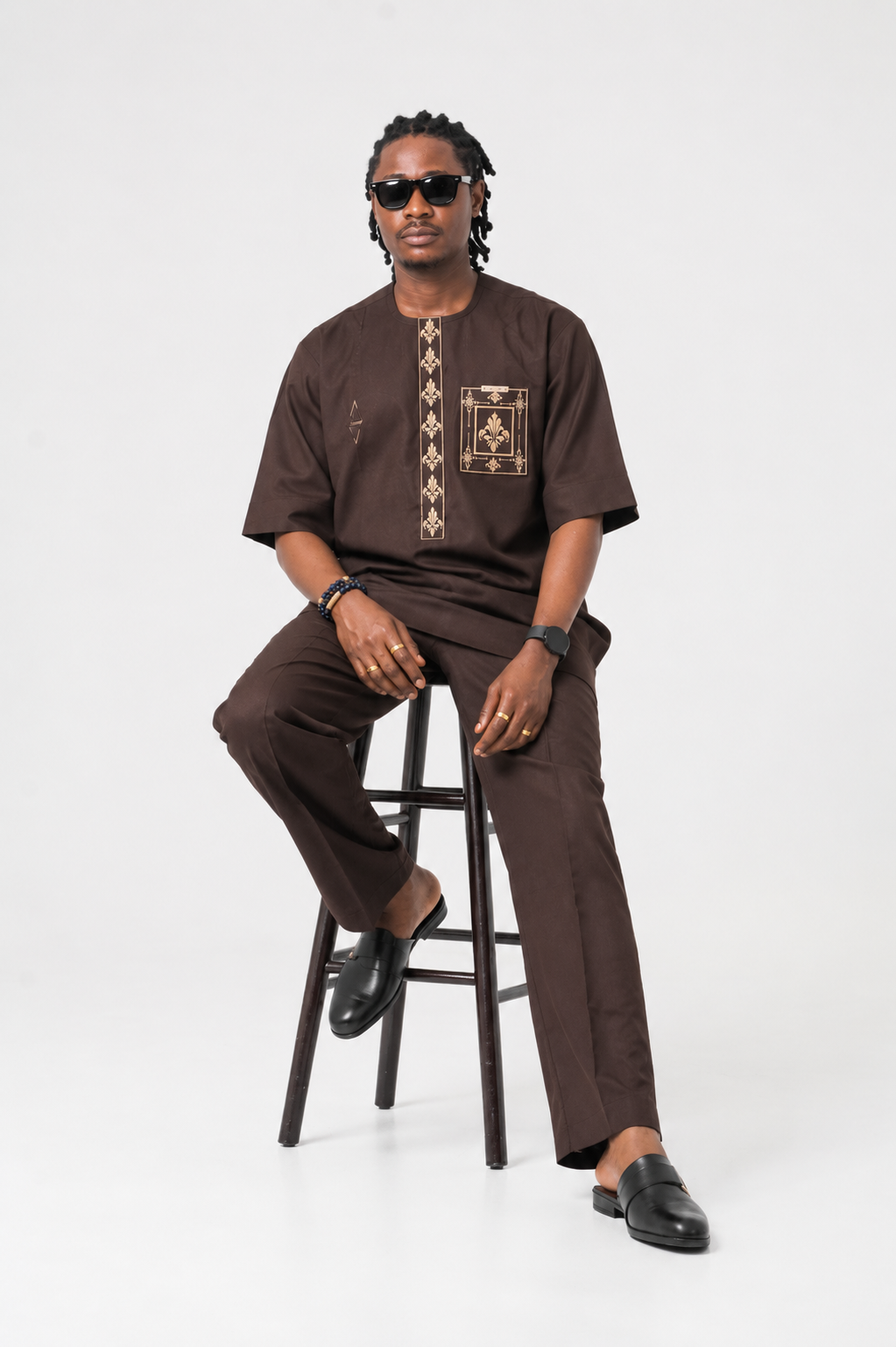 Man wearing a brown traditional outfit sitting on a stool against a white background