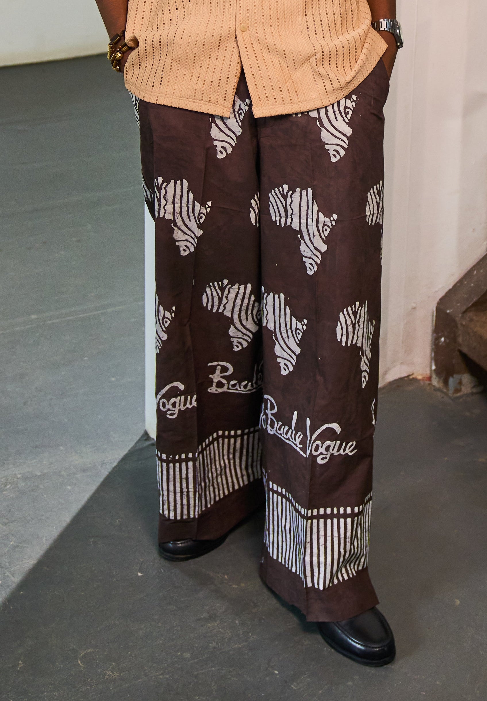 A man stands indoors wearing a light peach short-sleeve button-up shirt and dark brown wide-leg trousers with white graphic patterns and text. The room has a white wall, a colorful painting, a staircase, and a gray floor.