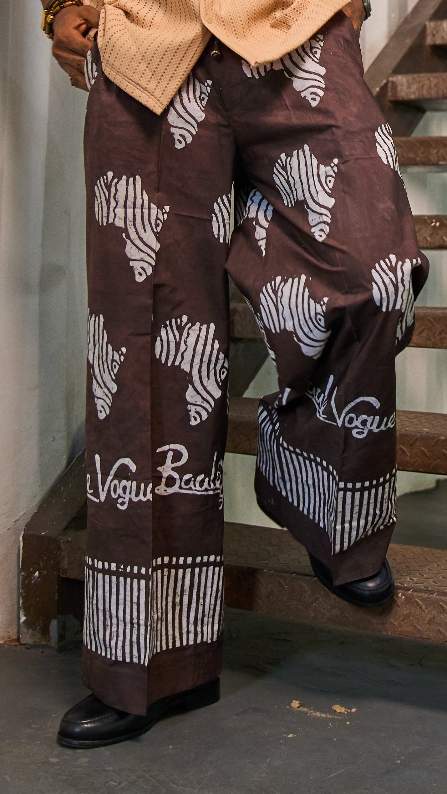 A man wearing a beige short-sleeve button-up shirt and brown wide-leg pants with white zebra and script patterns, standing on a wooden staircase in front of a white paneled wall.