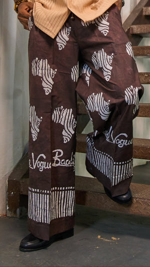 A man wearing a beige short-sleeve button-up shirt and brown wide-leg pants with white zebra and script patterns, standing on a wooden staircase in front of a white paneled wall.
