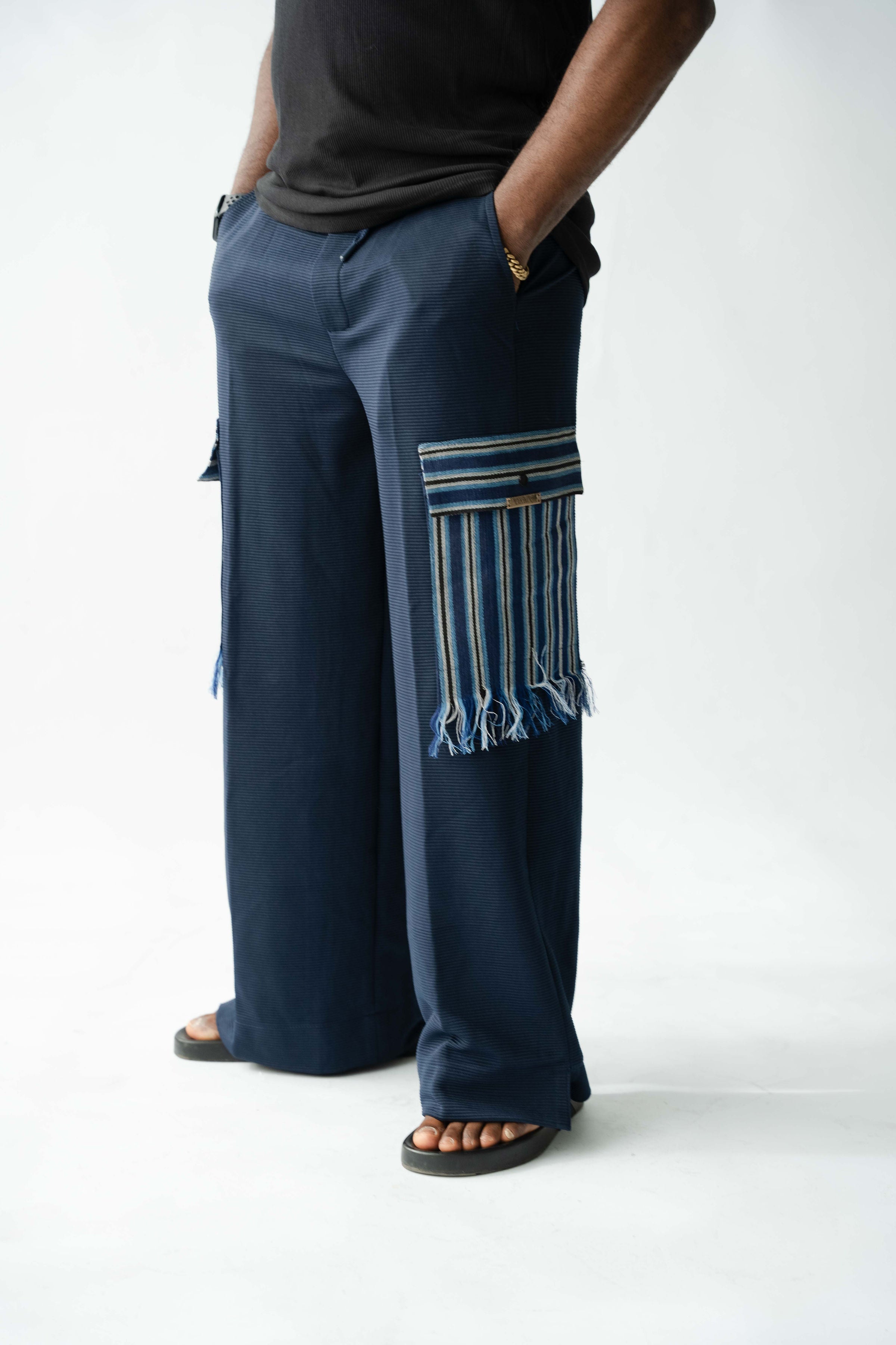 Navy wide-leg men's cargo trousers with handwoven Aso Oke pocket detail and fringe trim, side profile view.