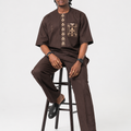 Man wearing a brown traditional outfit sitting on a stool against a white background